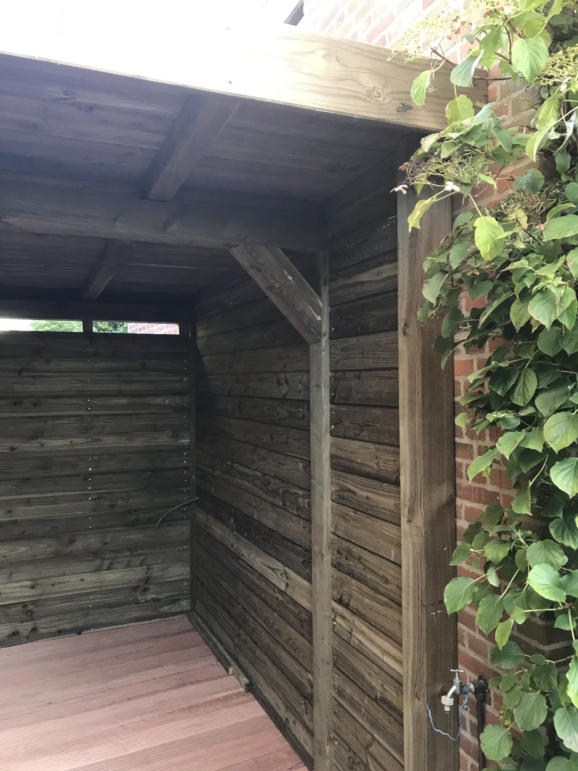 Houten schuur met horizontale lambrisering, open aan de linkerkant. Klimplanten tegen de bakstenen muur aan de rechterkant.