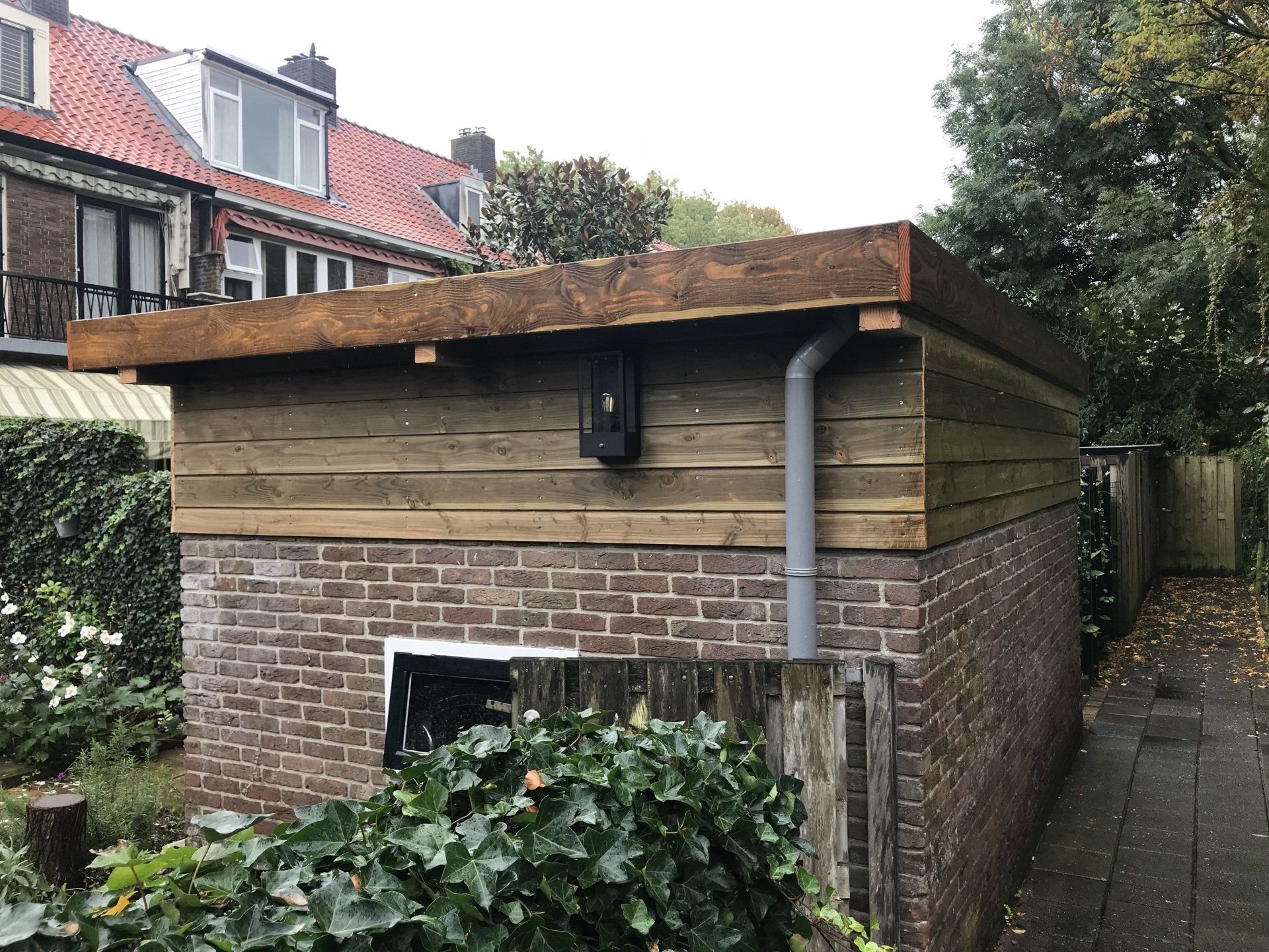 Een schuur van baksteen en hout met een plat dak, een grijze regenpijp en een zwarte elektriciteitskast.