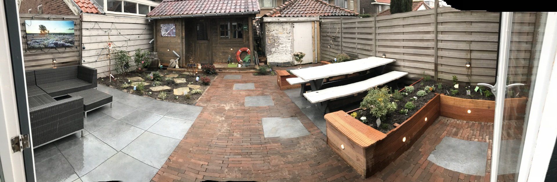 Achtertuin met terras, houten schutting, schuur, zitgedeelte, picknicktafel en verhoogde plantenbedden.