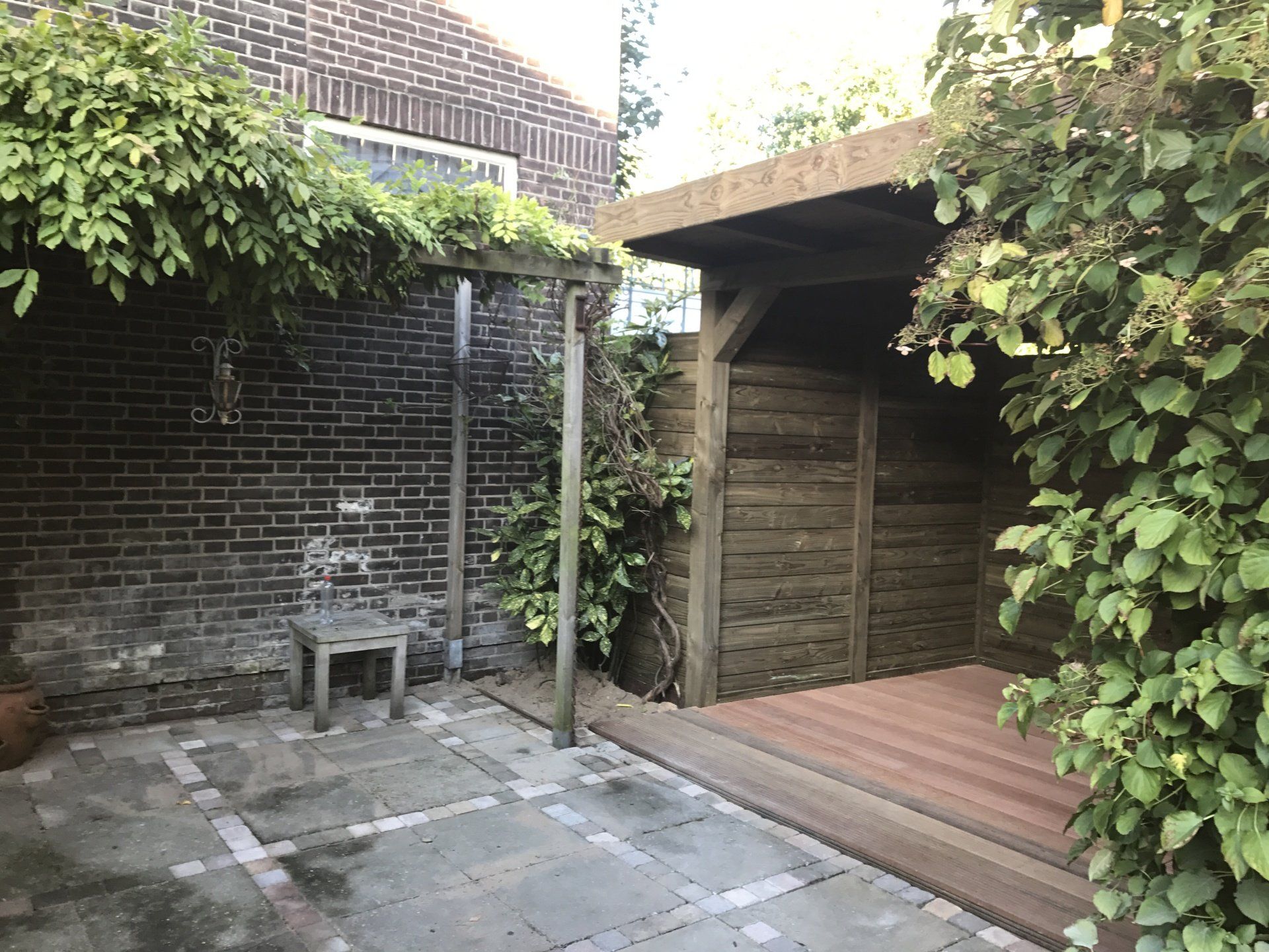 Een stenen terras met een houten schuur en klimplanten. Onder een houten pergola staat een stoel.