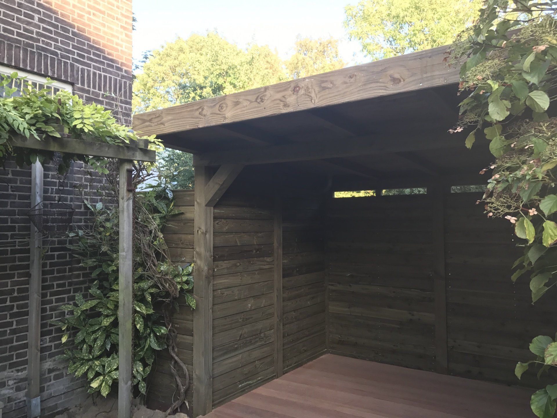 Een houten carport, aangebouwd aan een bakstenen gebouw, is omgeven door groen.