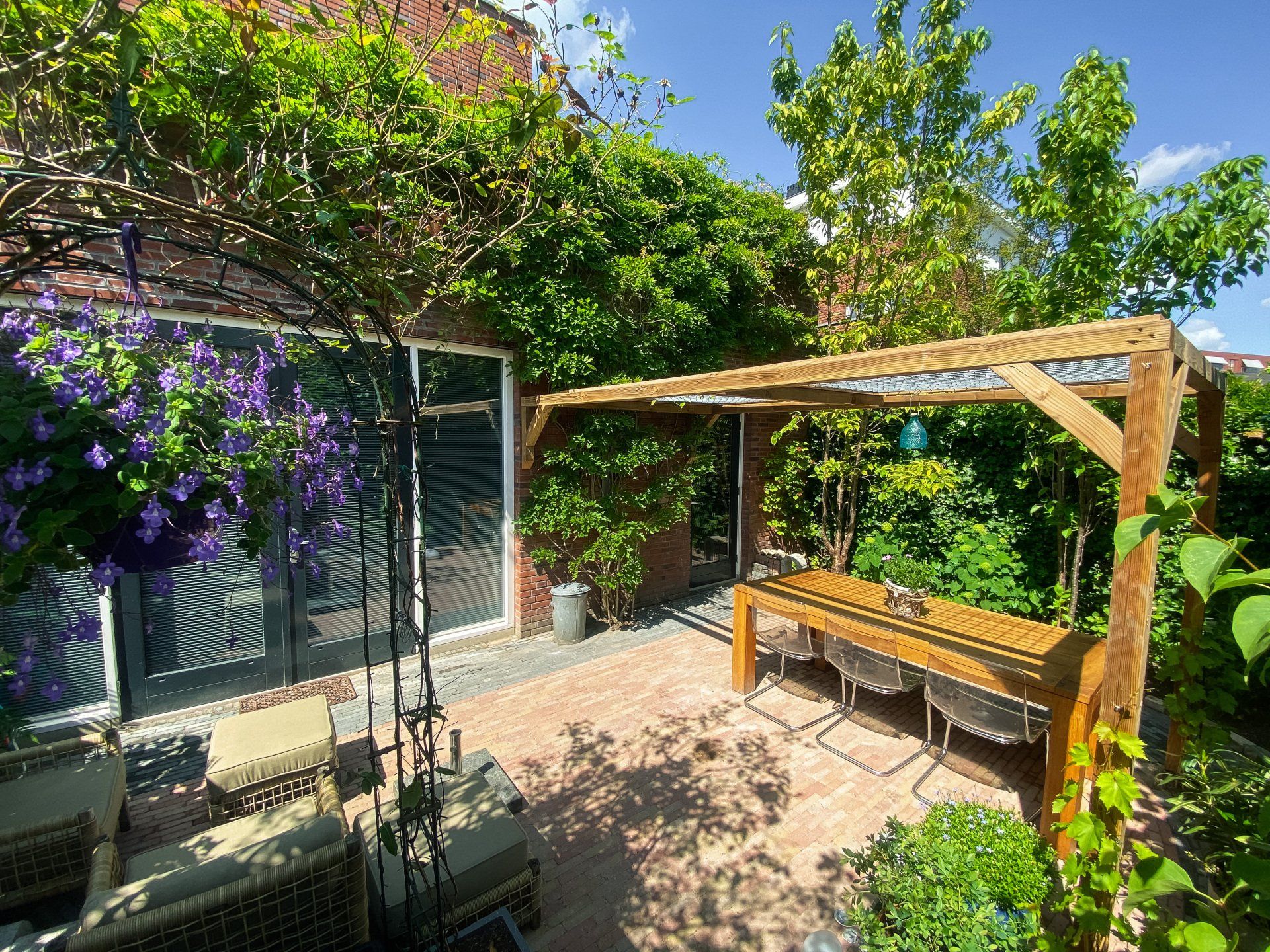 Een zonnig terras in de achtertuin met een houten pergola, bakstenen vloer en weelderig groen.