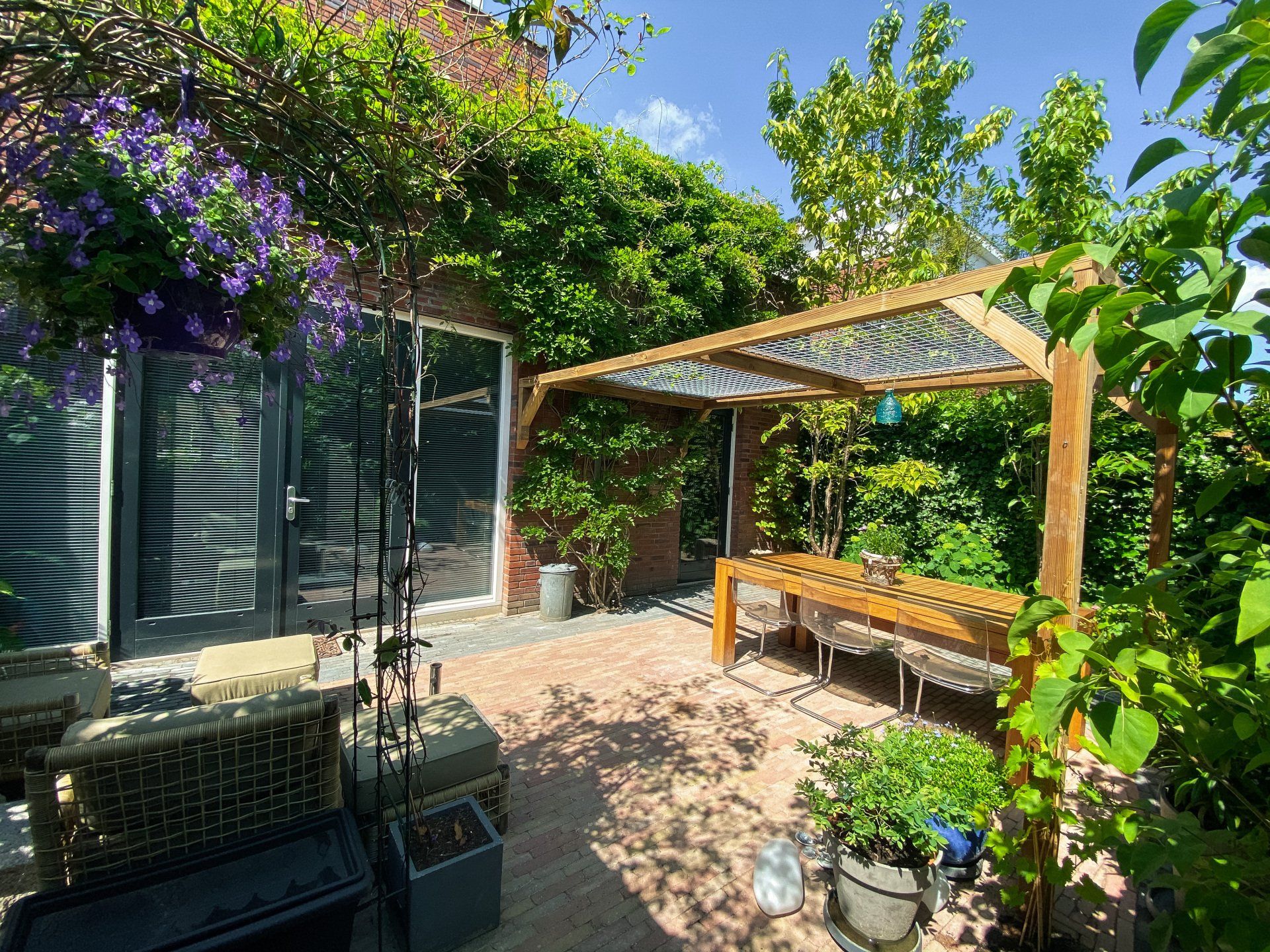 Buitenpatio met houten pergola boven een tafel. Bakstenen, planten en klimplanten creëren een zonnige ruimte.