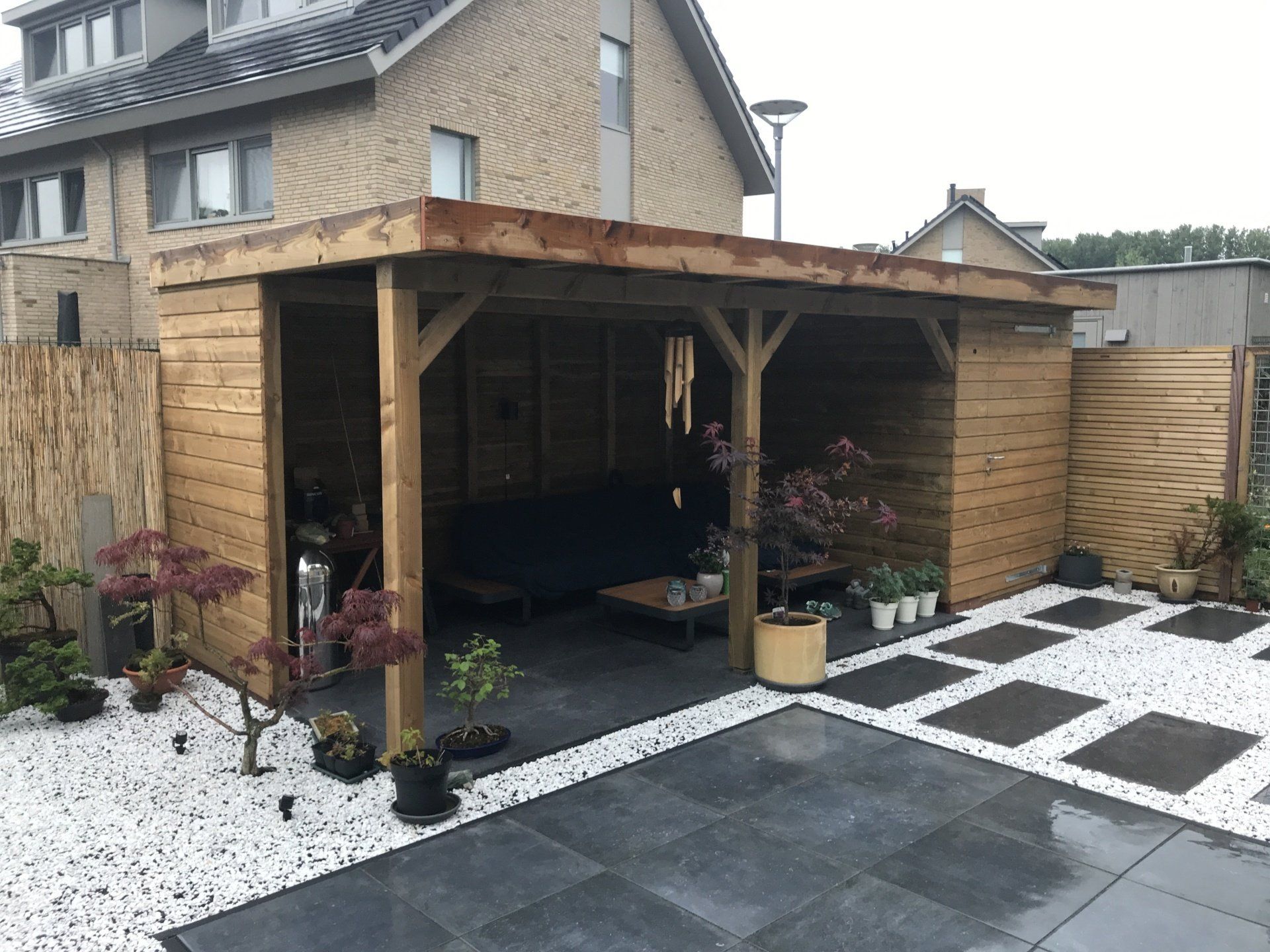 Houten terrasoverkapping met zitgedeelte, omgeven door planten, een stenen pad en witte kiezels.