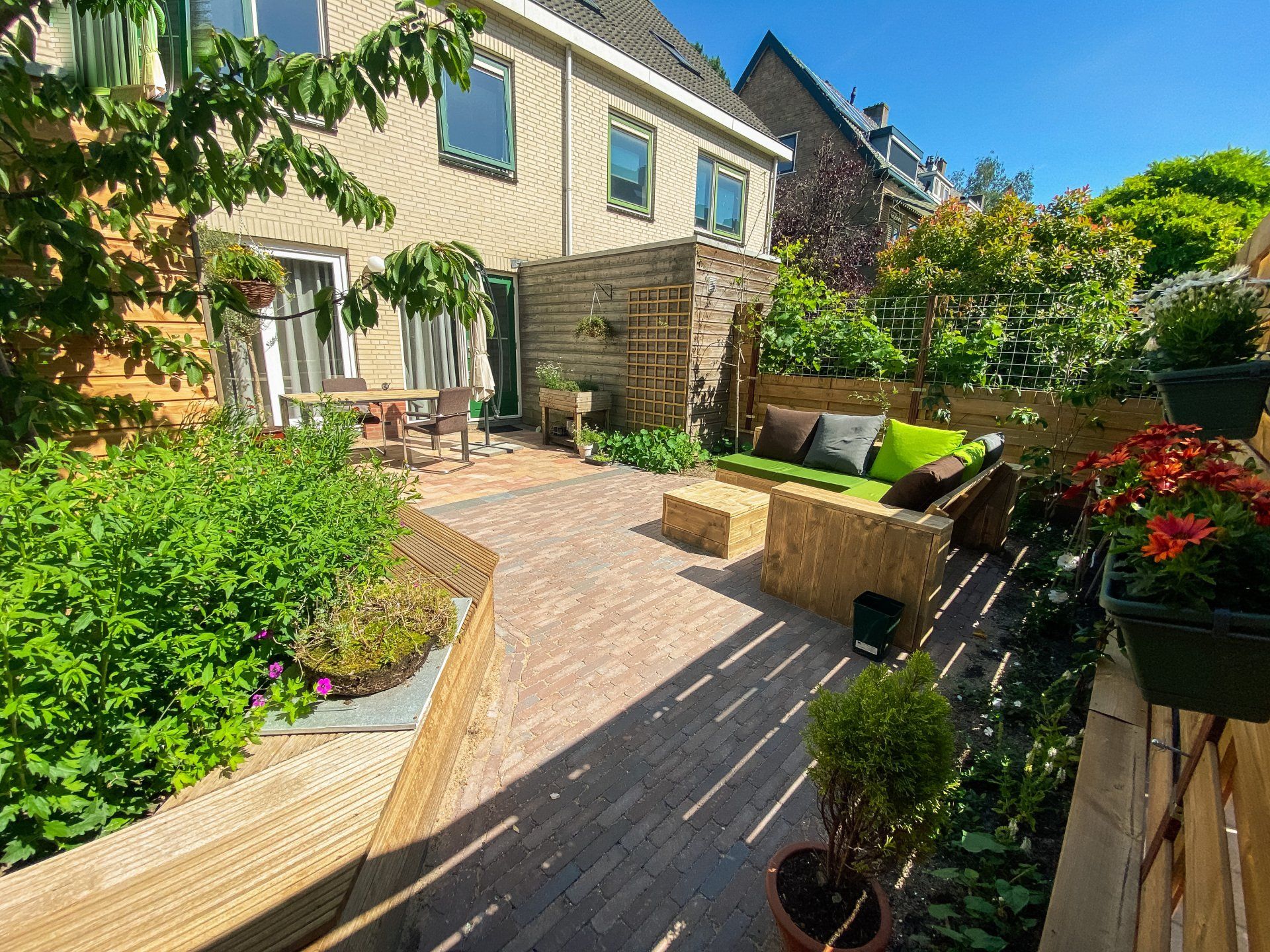 Gezellig terras met strobalenmeubilair, weelderige planten en een bakstenen pad. De zon stroomt naar binnen.