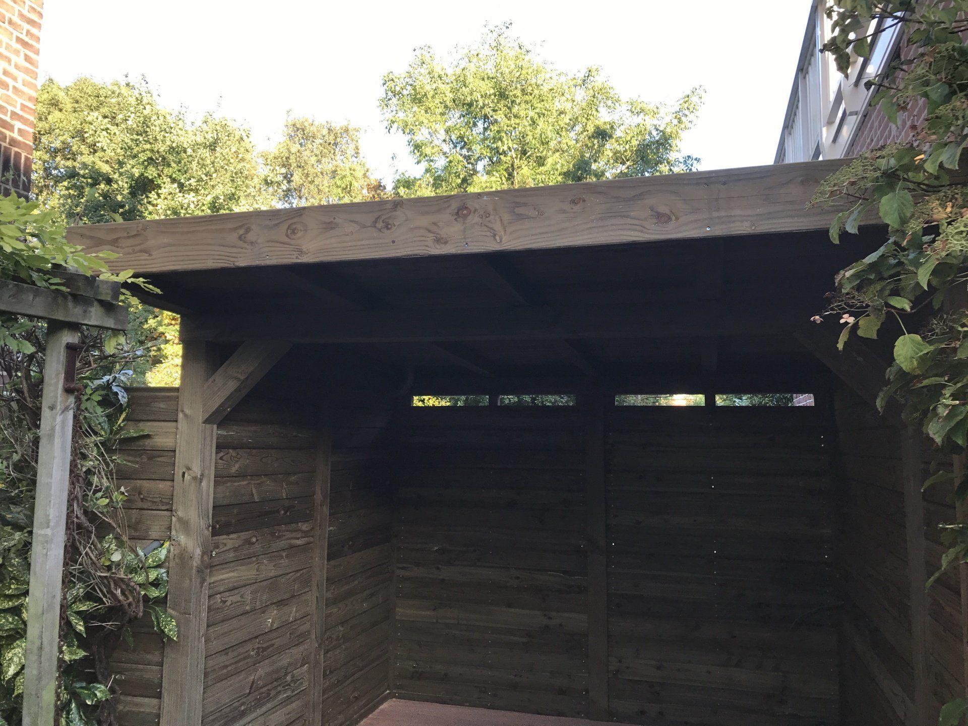 Houten schuur met een open voorkant en een plat dak, in een tuinomgeving.