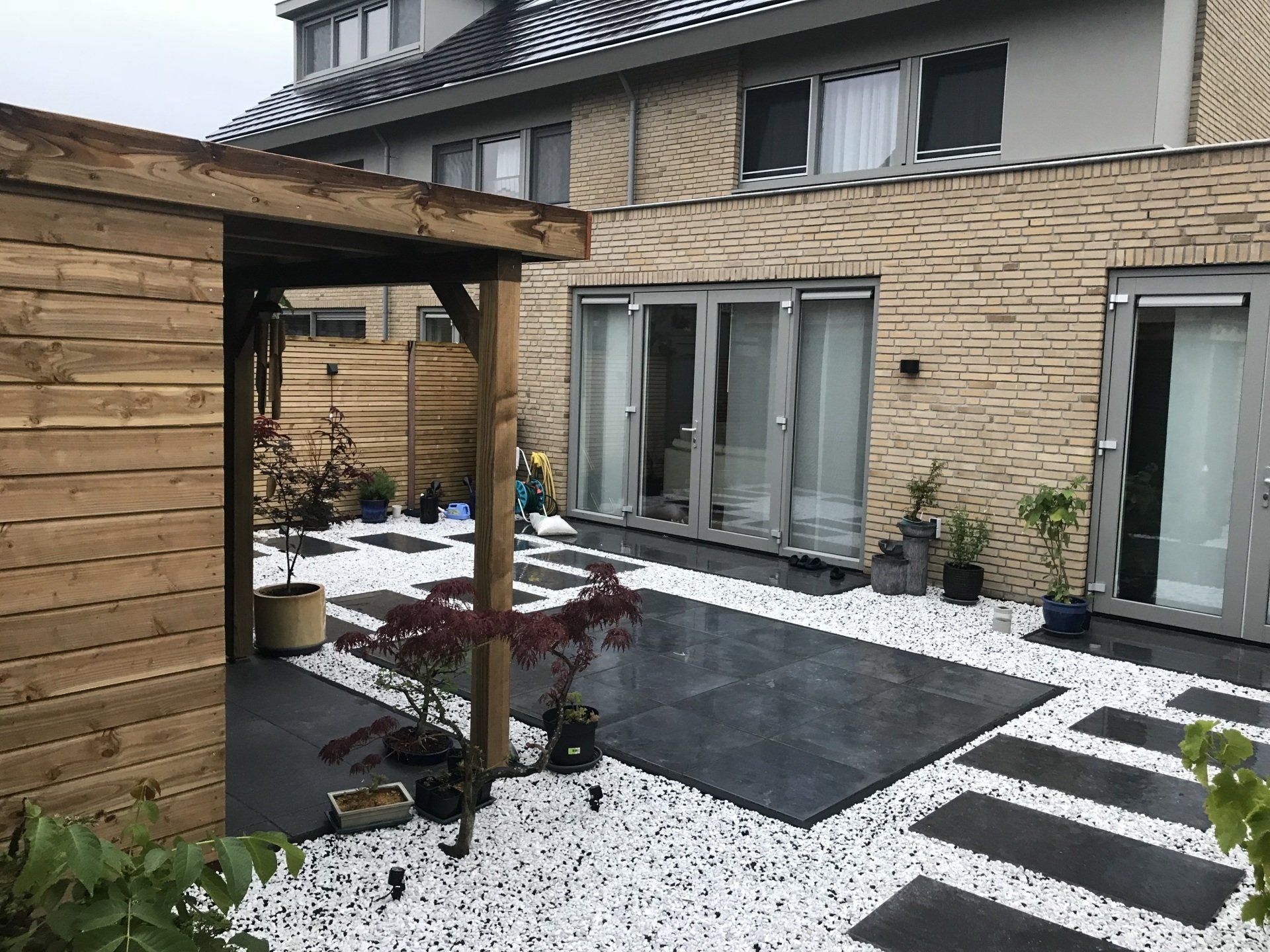 Terras met stenen bestrating, wit grind, houten pergola en een huis met glazen deuren.