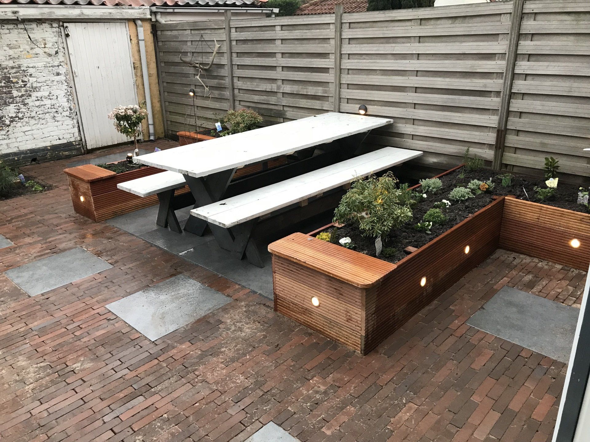 Achtertuin met stenen terras, picknicktafel en verhoogde plantenbakken. Houten schutting en de zijkant van een gebouw op de achtergrond.