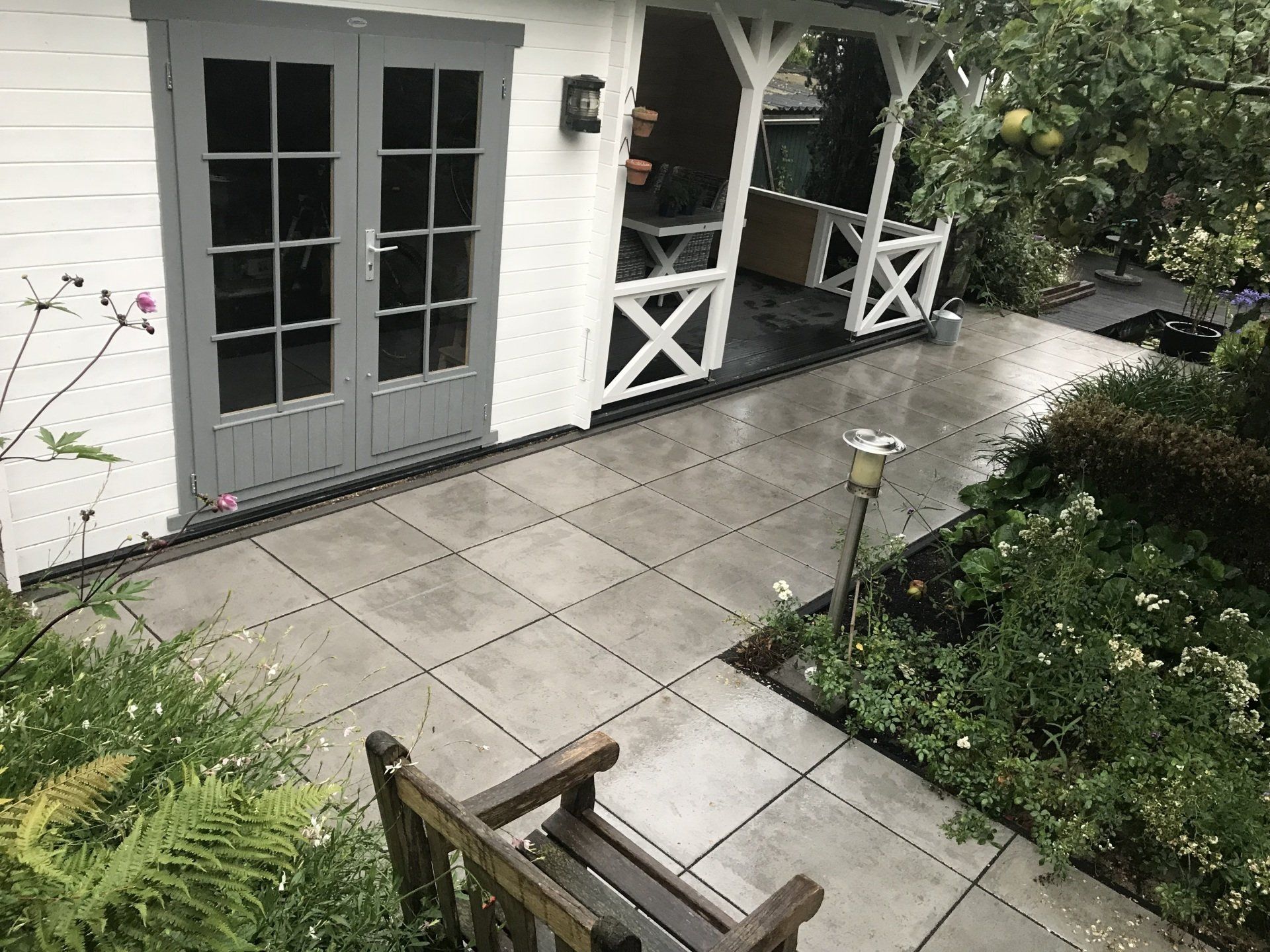 Grijze patio met een witte schuur, een houten bank en groene planten.
