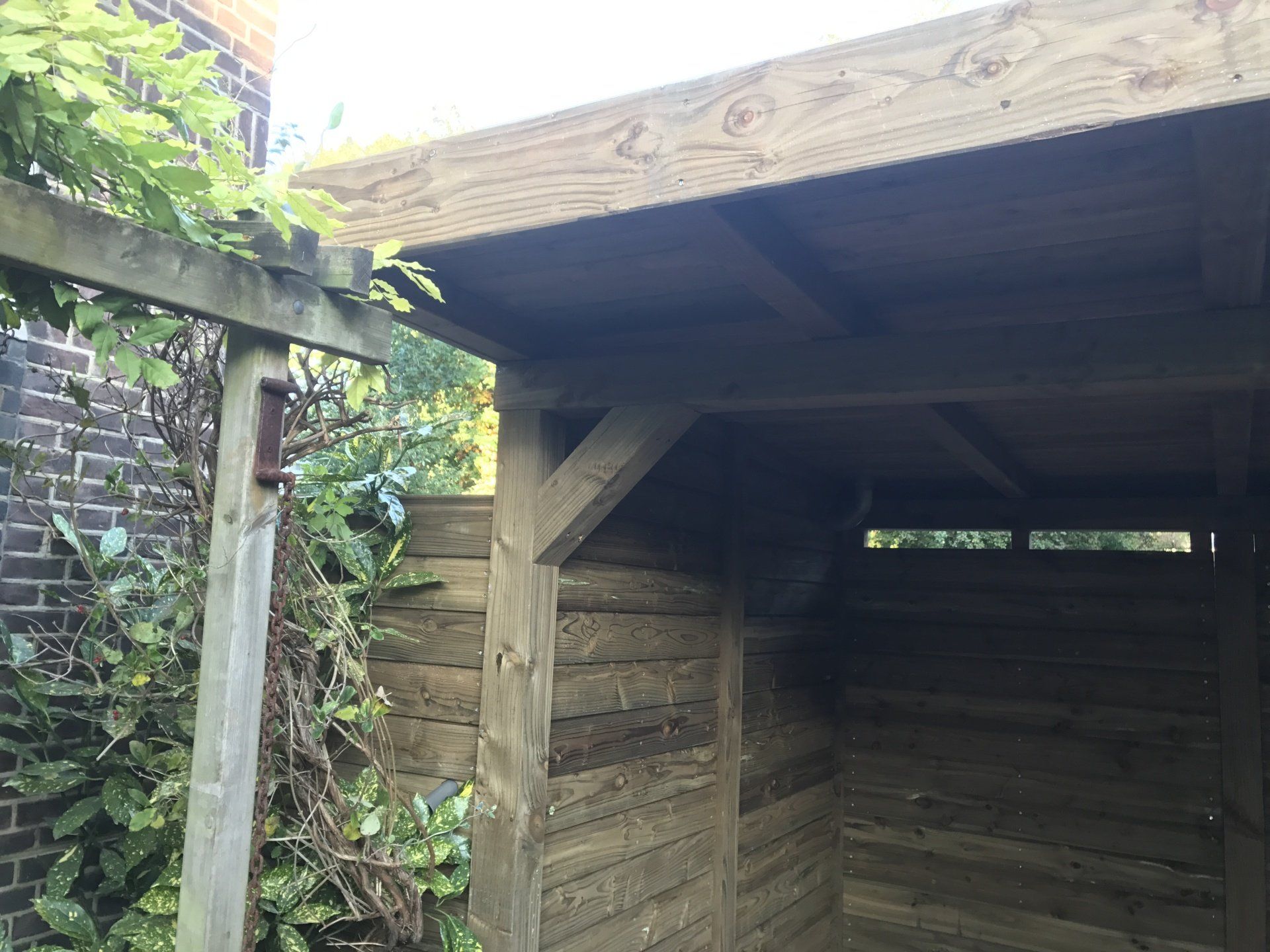 Houten schuur met overstekend dak in een tuin, gebouwd tegen een bakstenen muur.