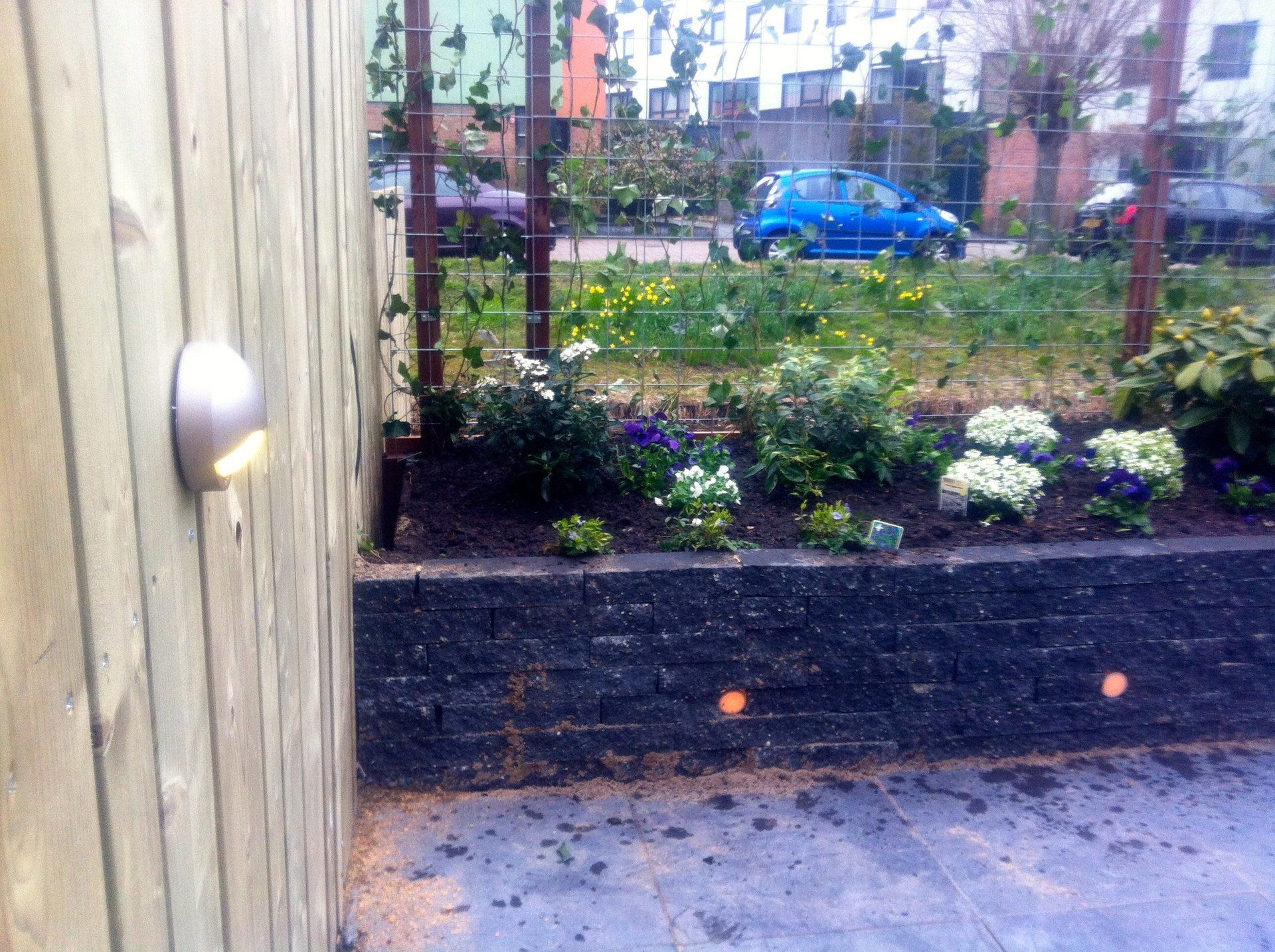 Houten schutting met ingebouwde verlichting, een bloembed met diverse bloemen en een blauwe auto.