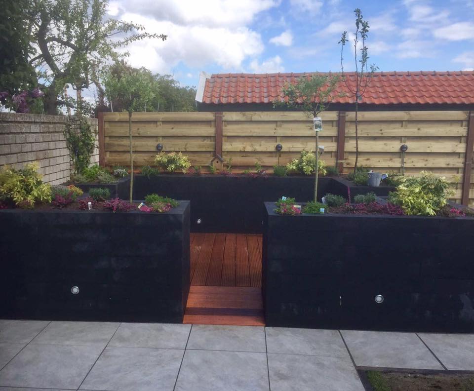 Verhoogde plantenbedden met een houten pad, tegen een houten schutting en een rood pannendak.