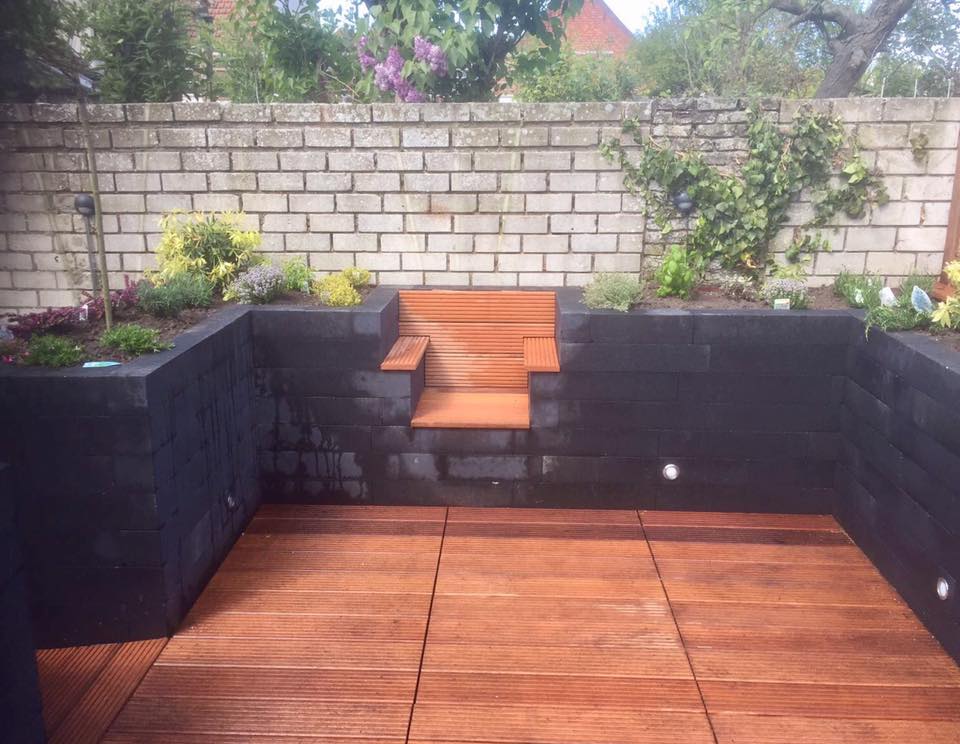 Terras in de achtertuin met houten vloer, zwarte keermuren en een terrasvormige tuin met een bakstenen achtergrond.