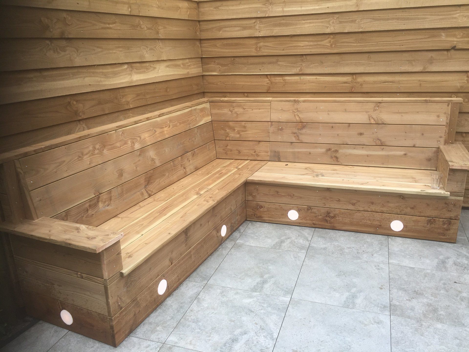 L-vormige houten buitenbank met ingebouwde verlichting, tegen een houten muur, op een grijs stenen terras.