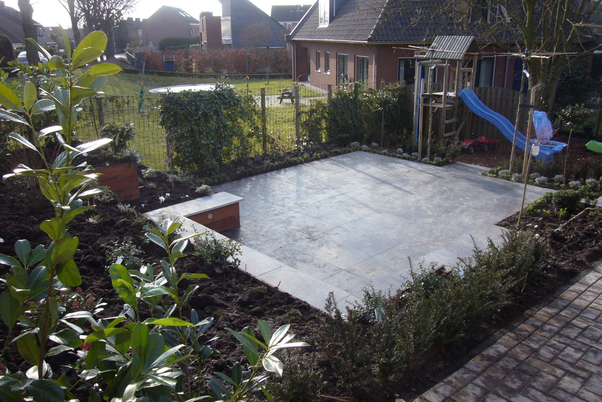 Terras met donkergrijze stenen, omgeven door groene struiken en een speeltoestel in de tuin.
