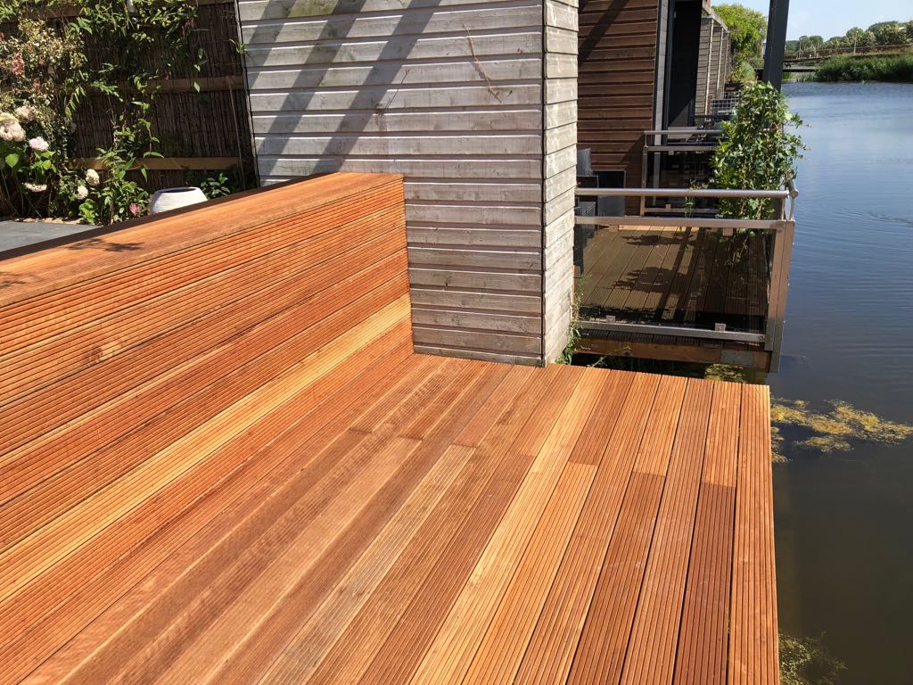 Houten terras op een woonboot, direct aan het water. Bruin hout, een metalen reling en een deel van een houten huis zijn zichtbaar.