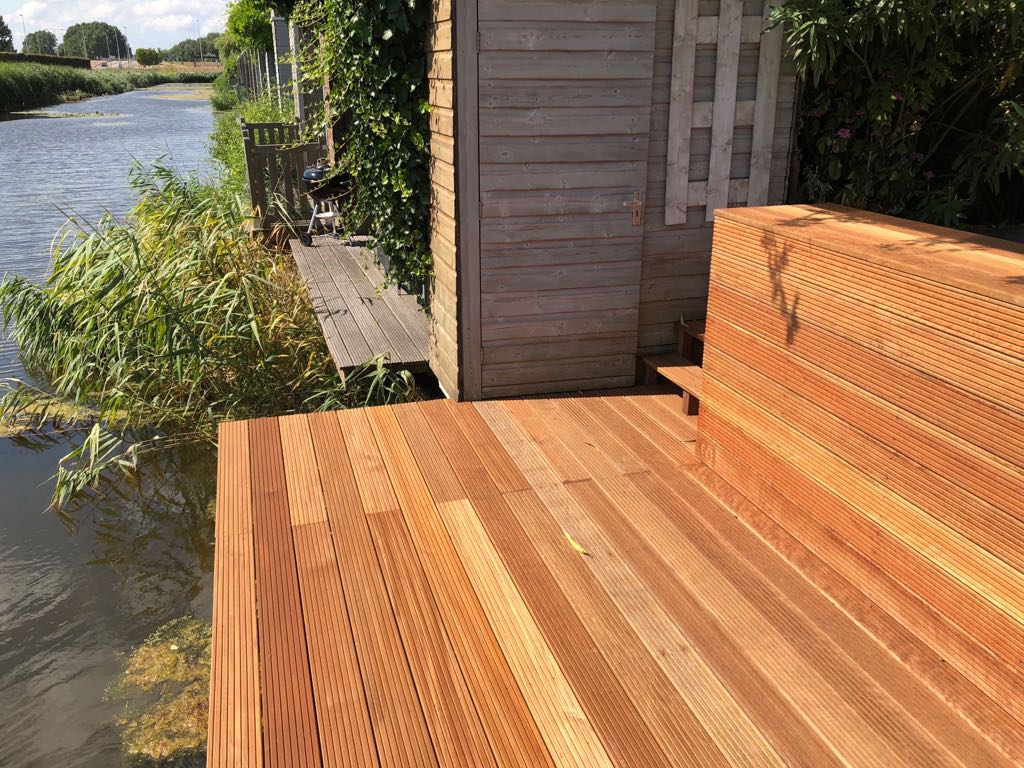Houten terras en bankje aan een kanaal. Schuur en begroeiing op de achtergrond. Fel daglicht.
