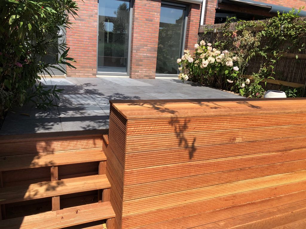 Houten terras met trappen, dat leidt naar een grijs terras bij een bakstenen gebouw met glazen deuren.