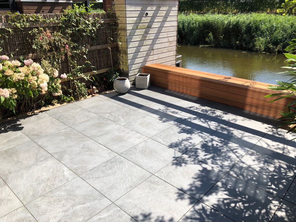 Terras met grijze stenen tegels, houten bank en een kanaal op de achtergrond.