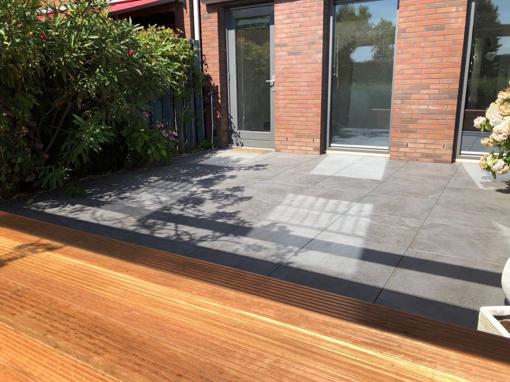 Terras met donkergrijze tegels, een houten vlonder en een bakstenen gebouw met glazen deuren, zonnige dag.