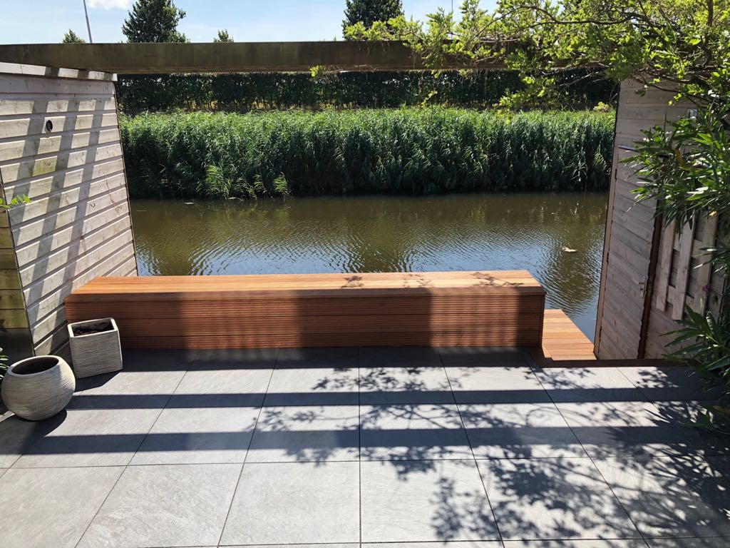 Een houten bank met uitzicht op een kanaal, geplaatst tussen grijze stenen pilaren en een donkere houten balk.