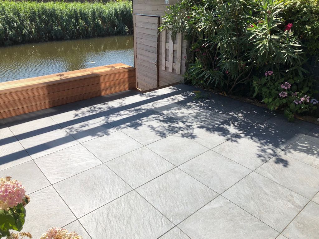 Terras met donkergrijze tegels aan het water, houten schutting en groen.