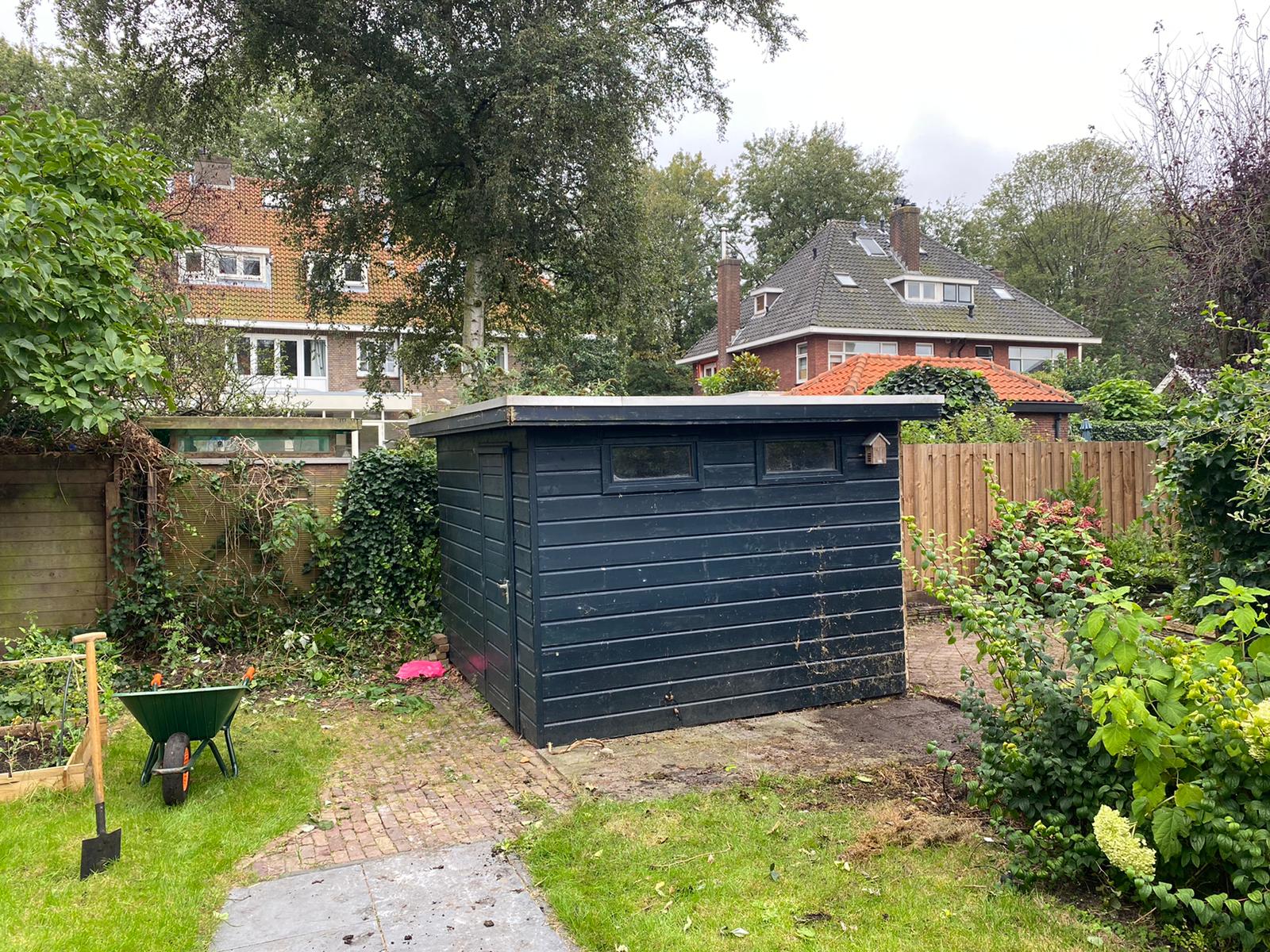 Zwarte houten schuur in een achtertuin met tuingereedschap, bomen en huizen op de achtergrond.