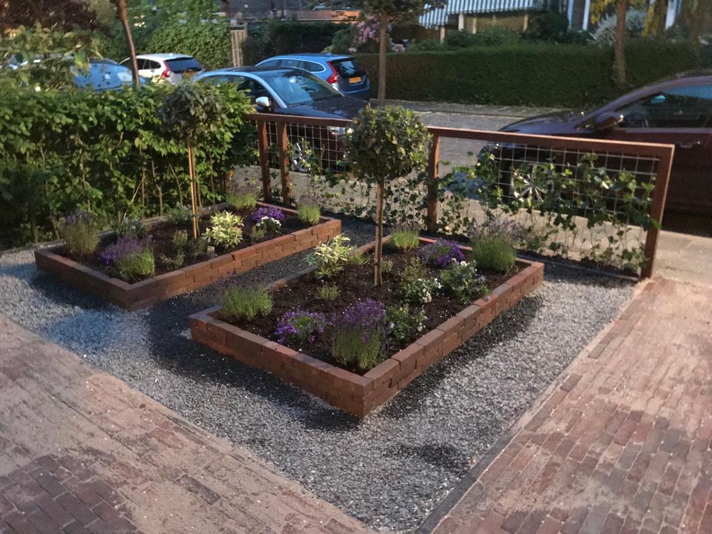 Verhoogde bloemperken met kleine boompjes, diverse bloemen en klimplanten, op een grindondergrond.