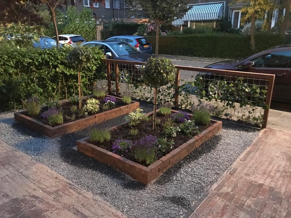 Twee verhoogde plantenbedden met planten en een grindlaag, omzoomd door hout, op een stoep, naast auto's.