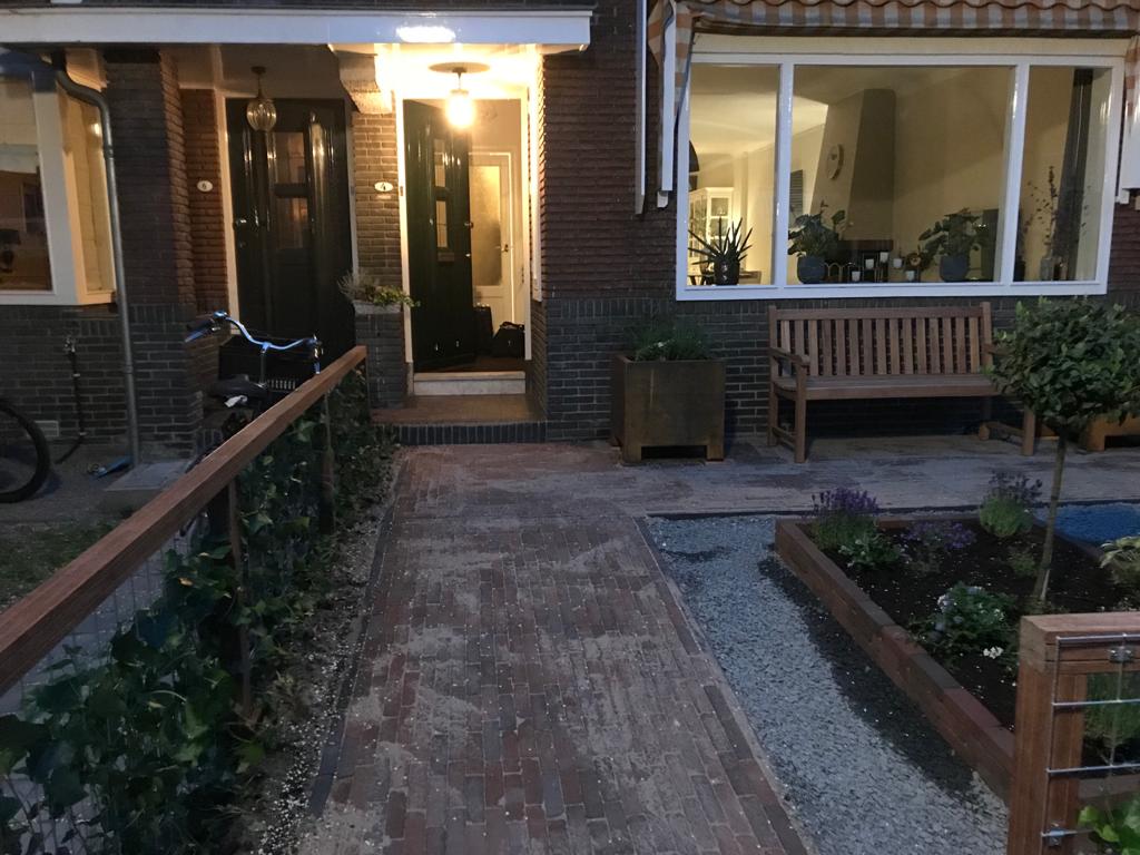 Een stenen pad leidt naar de ingang van een huis; het is voorzien van een bankje, bloembakken en een leuning.