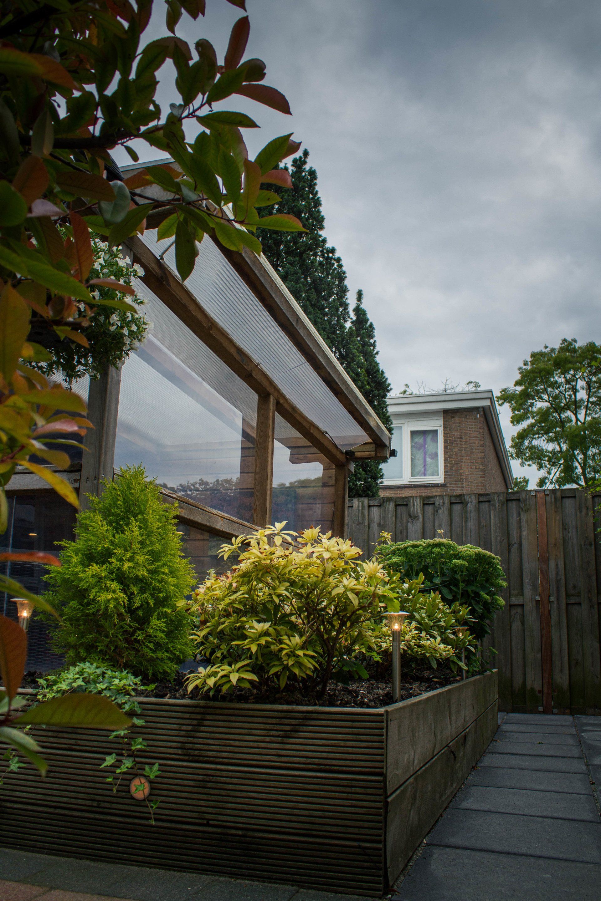 Verhoogd houten tuinbed met planten onder een transparant afdak, in een achtertuin.