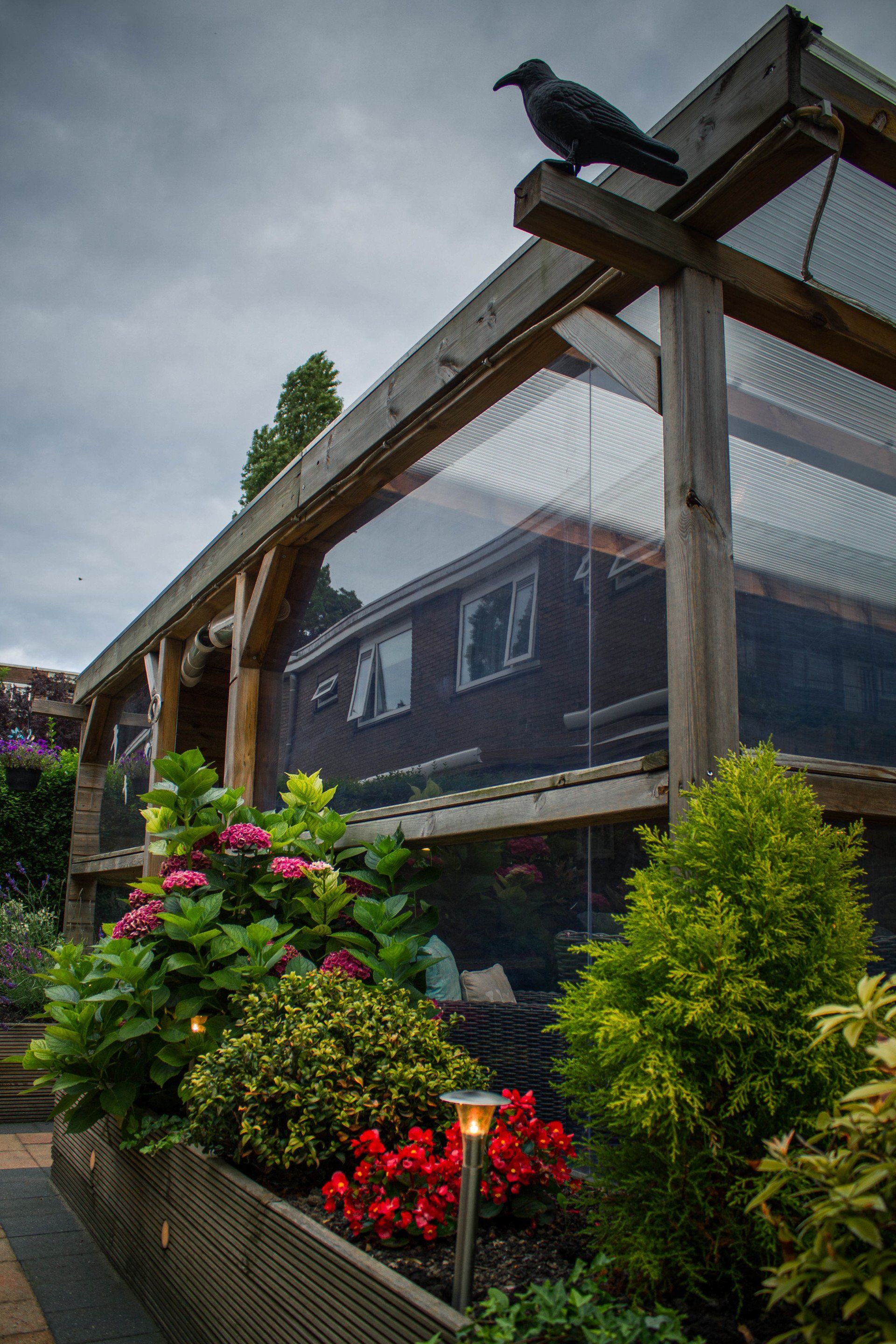 Houten volière met gaas, bloemen en een kraaienbeeldje bovenop. Bewolkte lucht op de achtergrond.