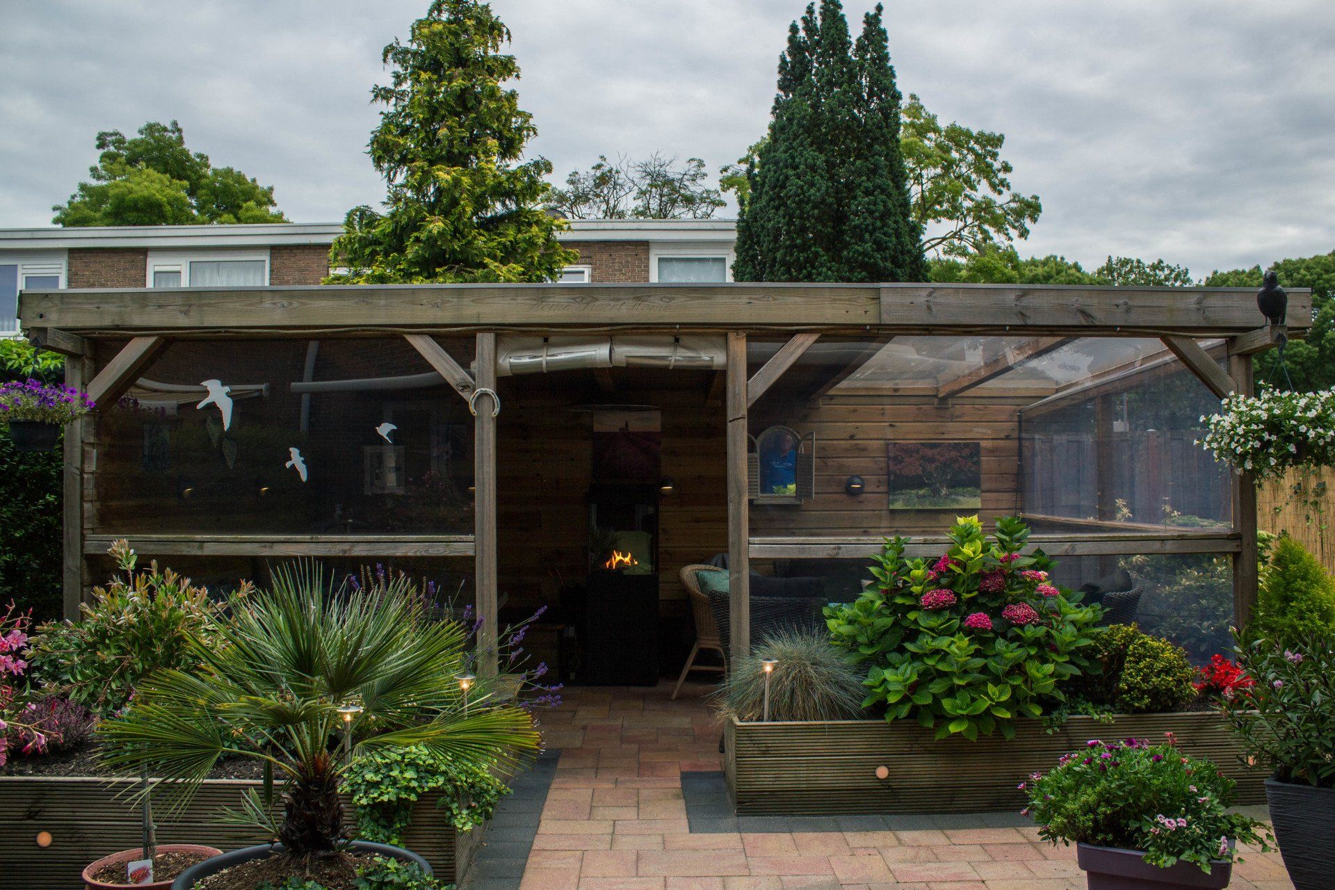 Houten volière met planten en bloemen, bakstenen pad, onder een bewolkte hemel.