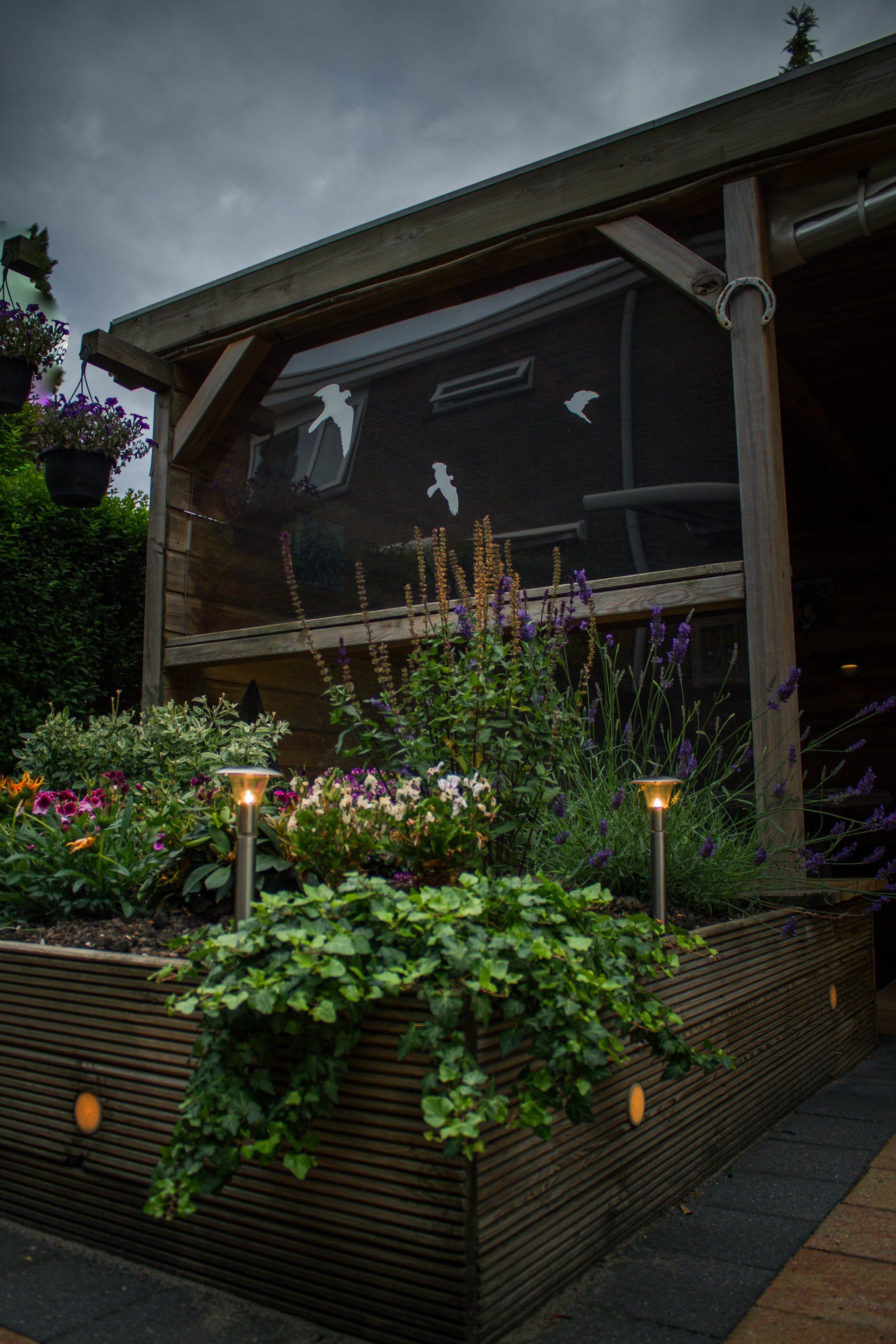 Verhoogd bloembed met bloemen en struiken, verlicht door lampen op grondniveau, naast een afgeschermde constructie onder een bewolkte hemel.