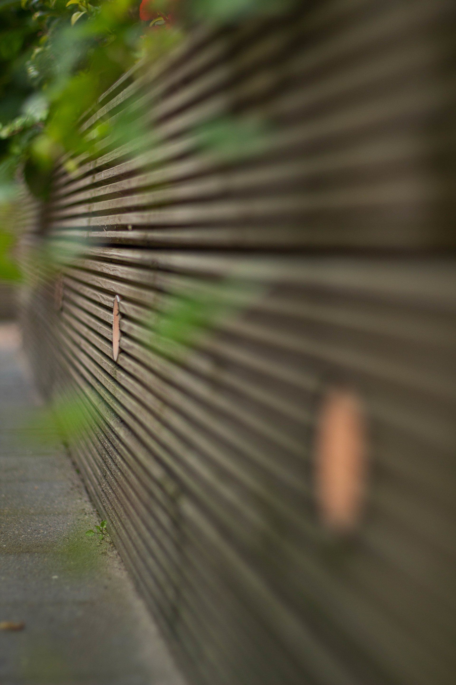 Houten schutting met horizontale latten, gedeeltelijk wazig, naast een trottoir en begroeiing.