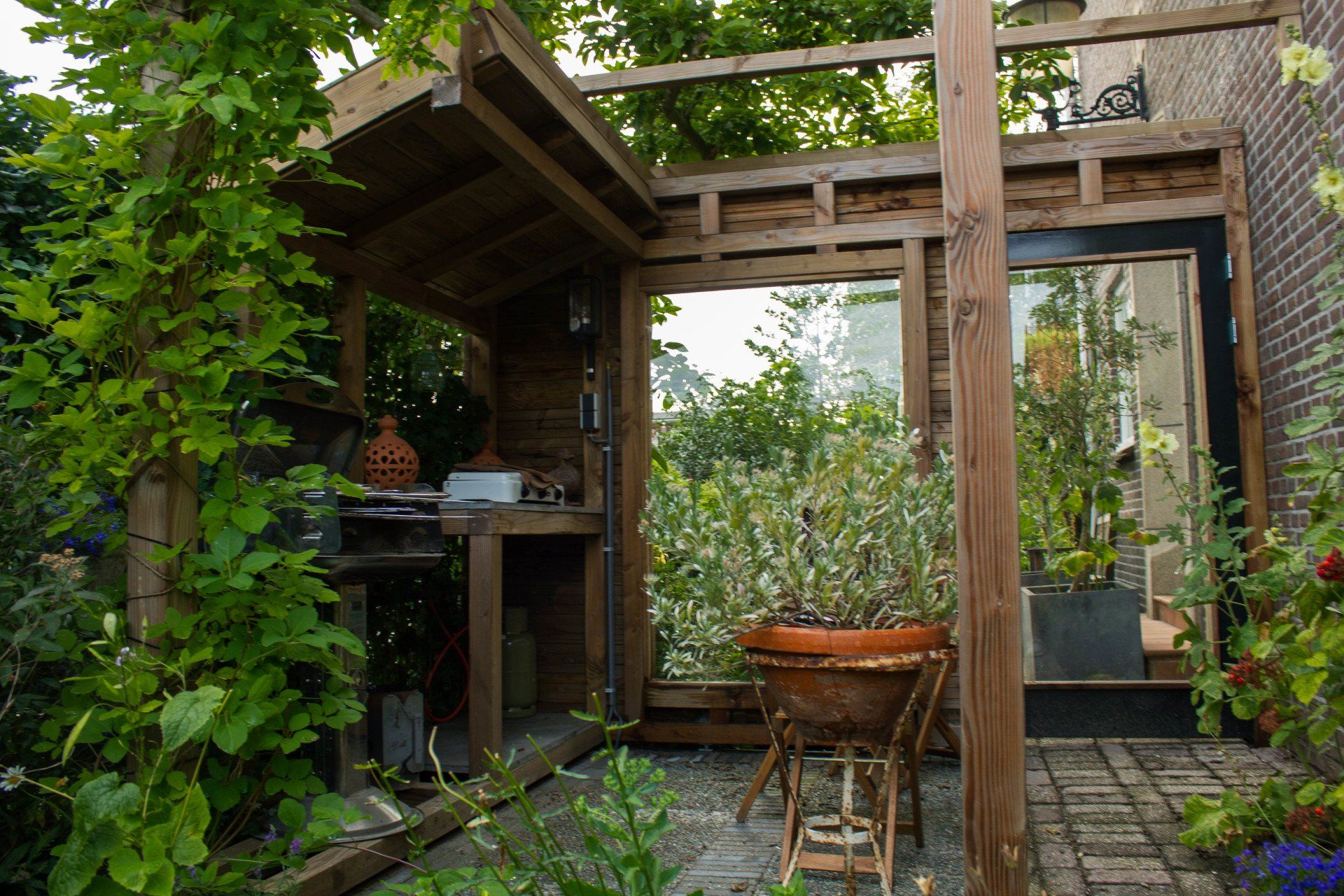 Houten buitenkeuken met grill en aanrecht, omgeven door planten en bakstenen.