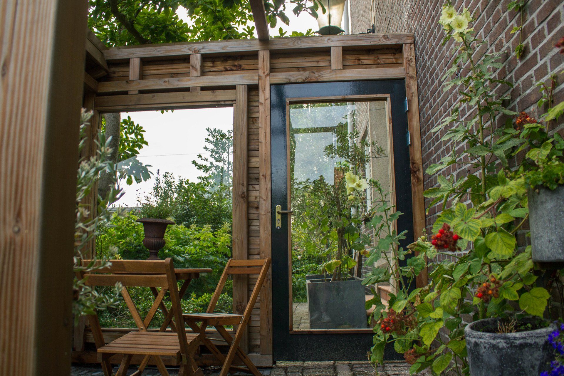 Houten terras met tafel en stoelen, een glazen deur met zwart frame en potplanten tegen een bakstenen muur.