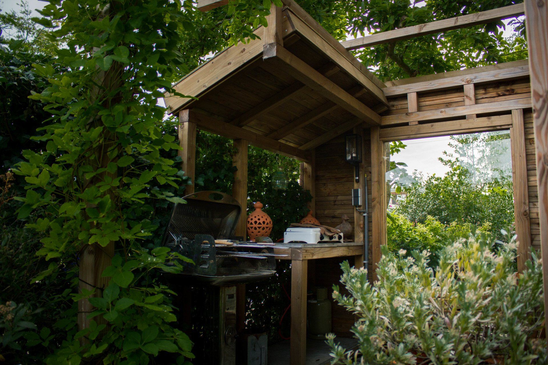 Houten buitenconstructie in een tuin, met een overdekte werkbank en decoratief aardewerk.