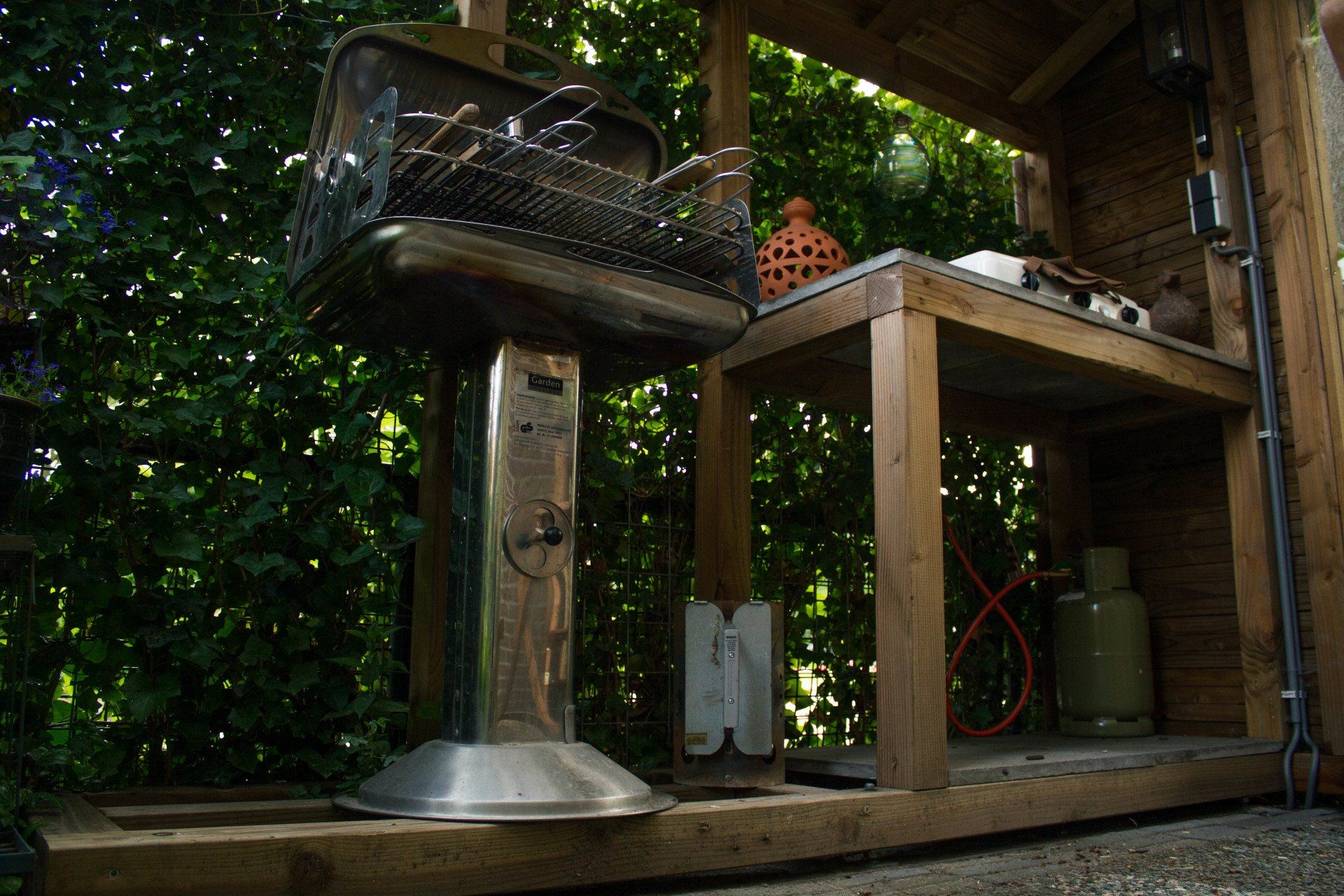 Buitenbarbecue op een houten onderstel met een opbergplank, omgeven door weelderig groen.