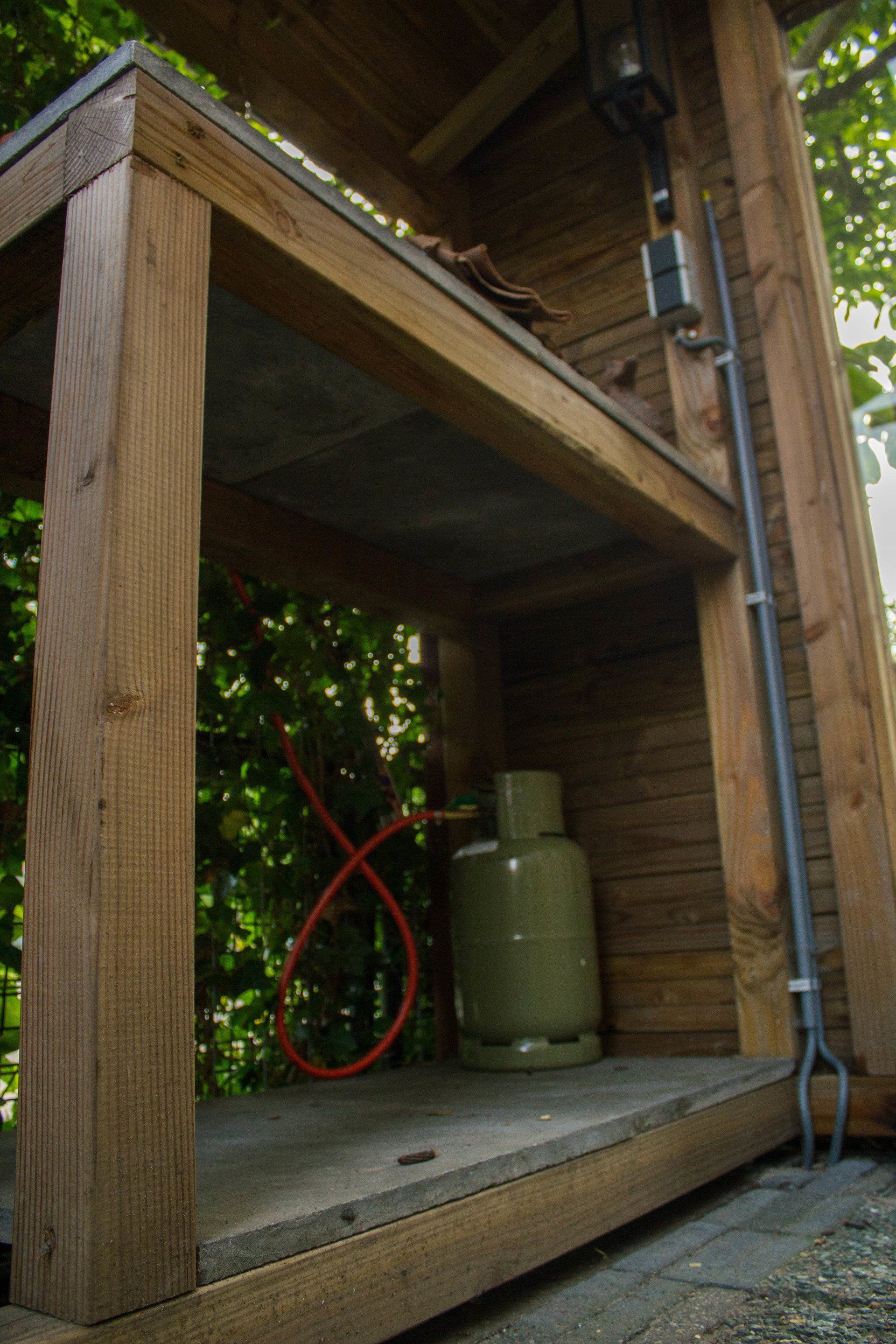 Houten constructie met een propaantank erin. Rode slang opgerold, metalen buis eraan bevestigd.