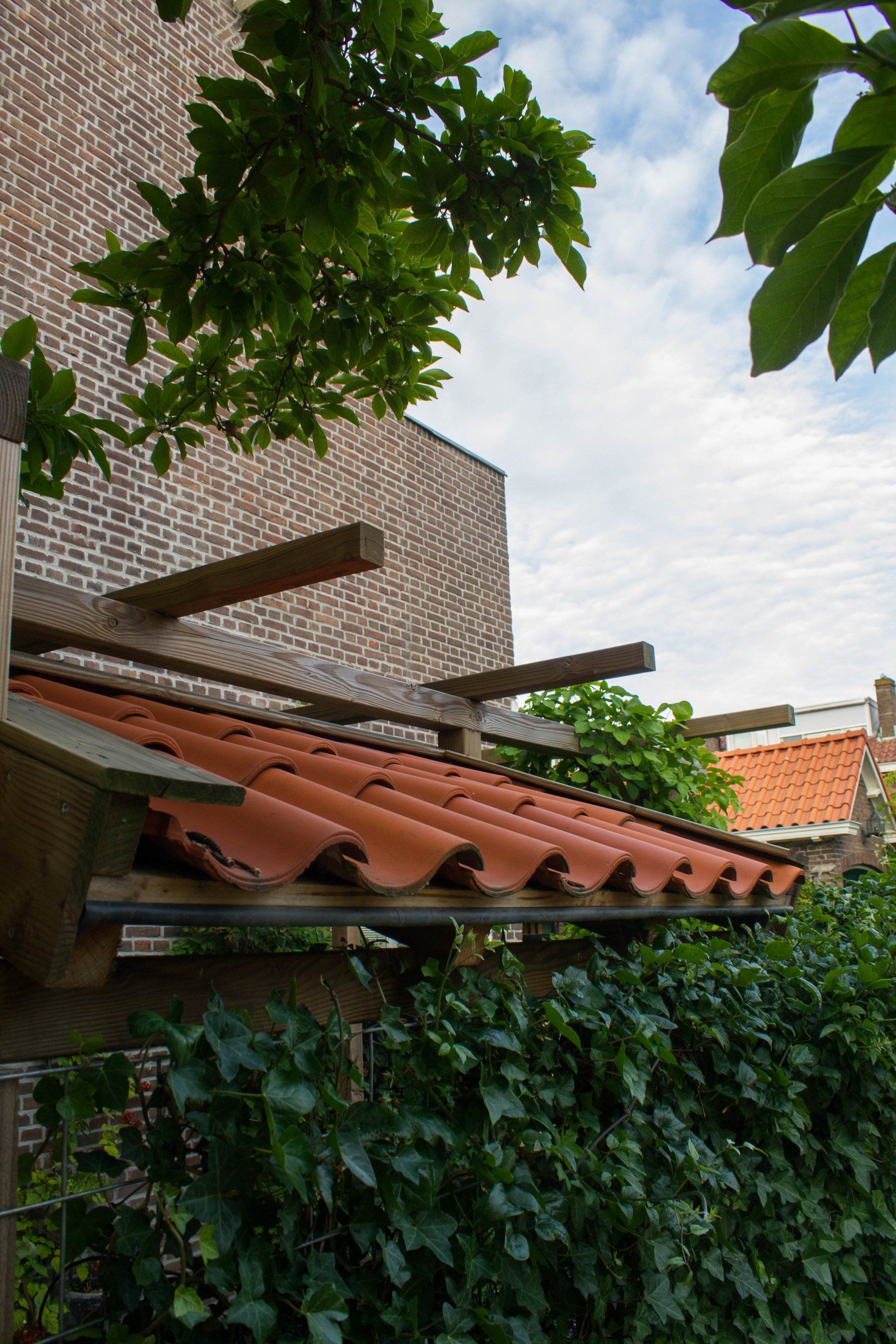 Een dak van terracotta dakpannen boven een groene muur, met een bakstenen gebouw en de lucht op de achtergrond.