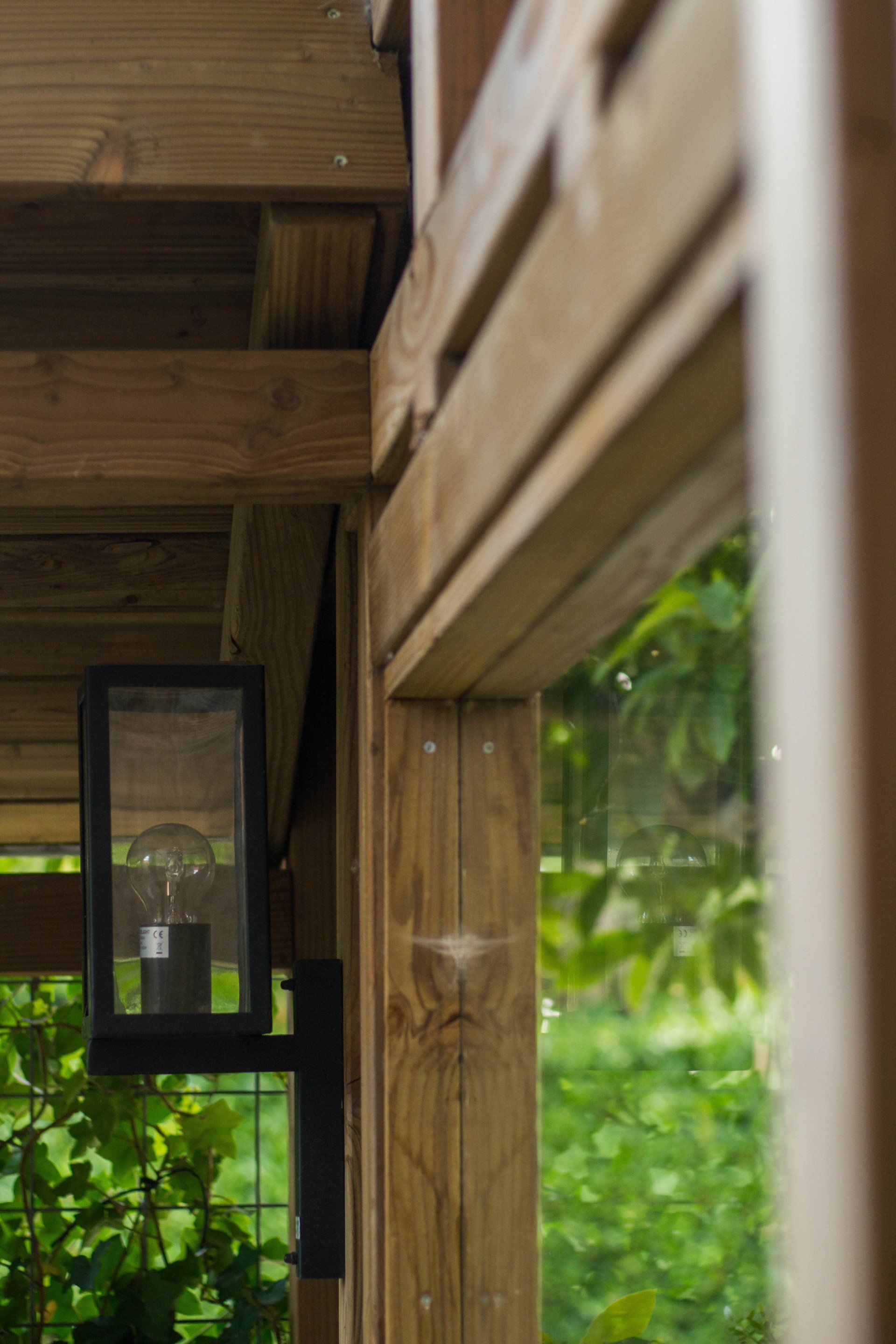 Houten verandahoek met een zwarte lantaarn en uitzicht op groen gebladerte door een raam.