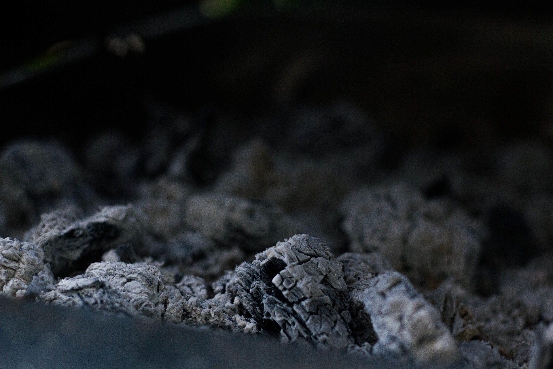 Close-up van grijze as en verkoold hout in een donkere omgeving, waarschijnlijk een barbecue of open haard.