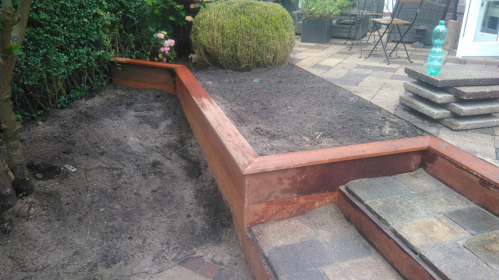 Verhoogd tuinbed met houten frame, gevuld met aarde. Terras op de achtergrond met planten en gestapelde bestrating.