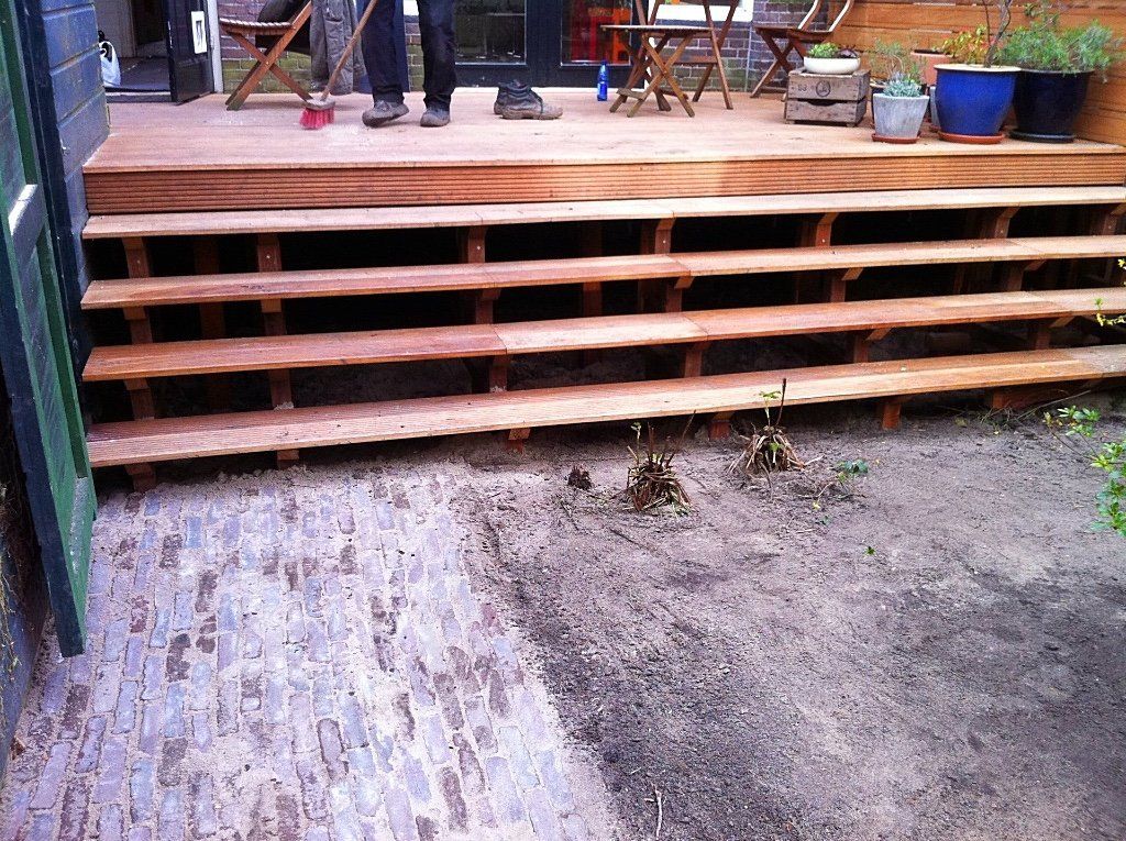 Houten terras met een aantal treden die naar beneden leiden naar een ondergrond van bakstenen en aarde; twee mensen staan ​​op het terras.
