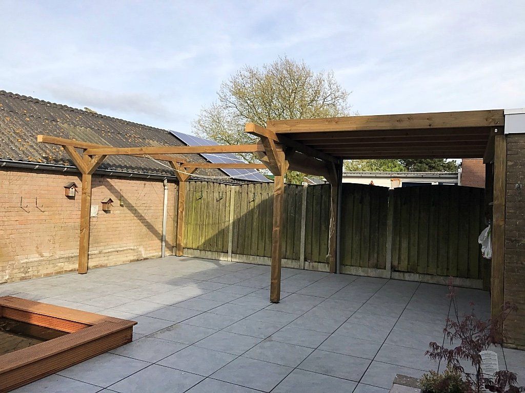 Houten pergola boven terras; bruine balken, grijze tegels, houten schutting, bakstenen muur, blauwe lucht.