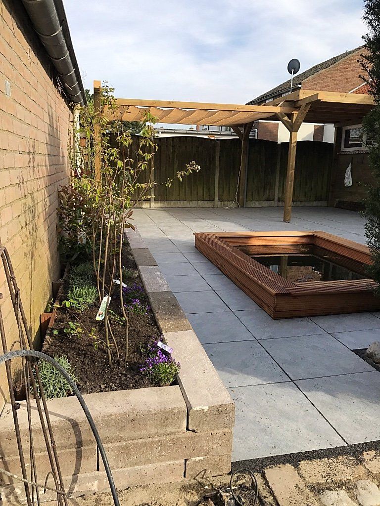 Achtertuin met een houten pergola, stenen terras, verhoogd tuinbed en een houten zandbak.