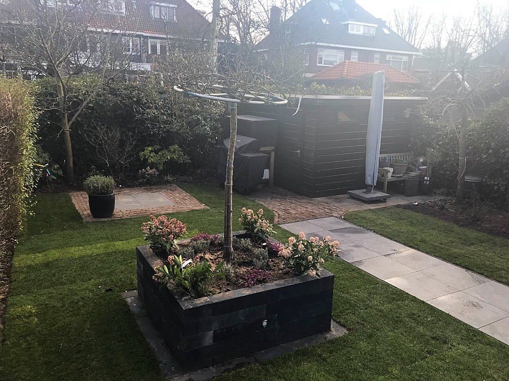 Tuin met gazon, bloemperken, bomen en een donker schuurtje. Zonlicht, diverse groensoorten, stenen pad.