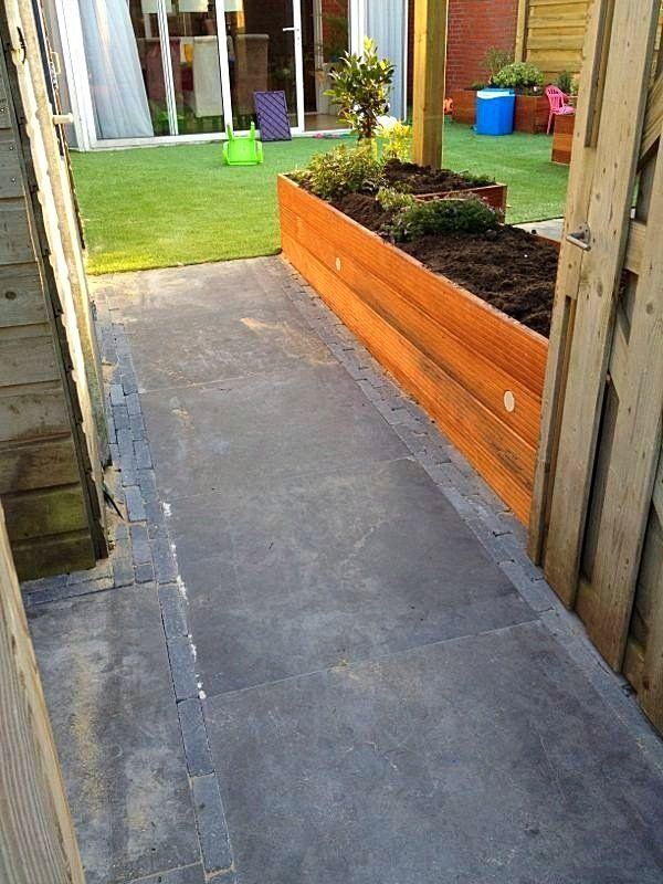 Een houten plantenbak naast een donkergrijs, geplaveid pad dat naar een grasveld leidt.