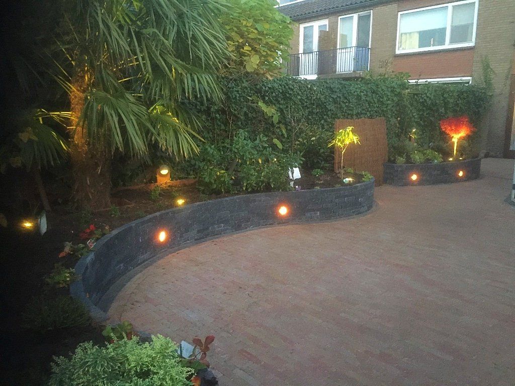 Een gebogen stenen muur met ingebouwde verlichting omlijst een aangelegde tuin met verlichte planten en een huis.